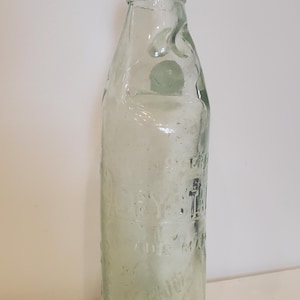 May include: A vintage clear glass bottle with a cork stopper. The bottle has a rounded shape and embossed text. The bottle is empty and has a light green tint, suggesting age. The bottle's height is approximately 20 cm.