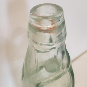 May include: Clear glass bottle with a cork stopper. The bottle has a rounded body and a narrow neck. The glass has a slight green tint. The bottle is empty. The bottle is likely vintage or antique.