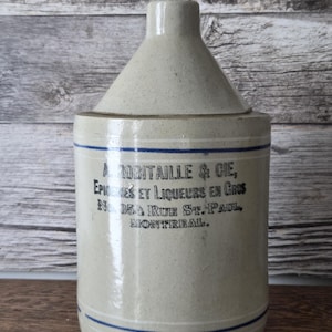May include: Vintage stoneware jug in a light beige hue, featuring blue stripes. The jug has a conical top and a cylindrical body. Text in black ink reads "A ROBITAILLE & CIE, ÉPICERIES ET LIQUEURS EN GROS, No. 354 RUE ST. PAUL, MONTREAL."