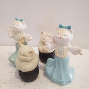 May include: Three vintage porcelain cat figurines with blue and white dresses and fans. The cats are in various poses and are holding fans. The figurines are on a white background.