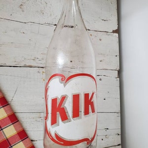 May include: A clear glass bottle with a red and white label that says "KIK" on it. The bottle is hanging on a white wooden wall.