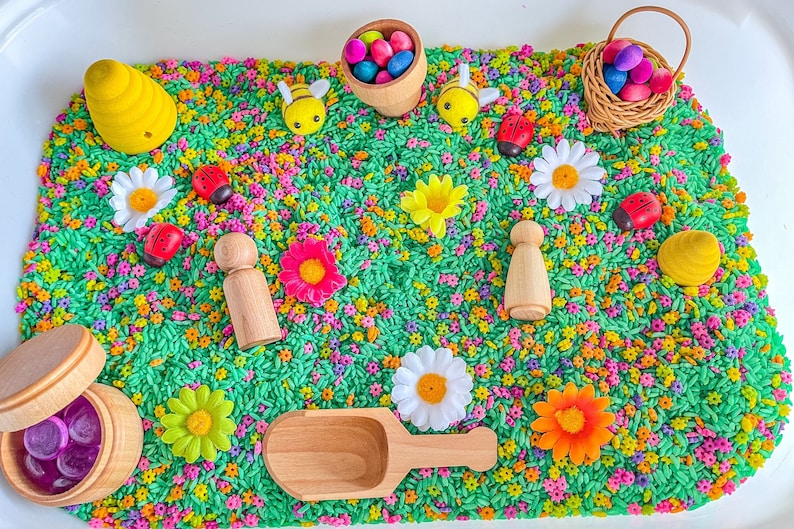 May include: A sensory bin filled with green rice and colorful flowers, ladybugs, bees, and wooden figures. There are also two wooden baskets, one filled with purple gems and the other with colorful eggs.