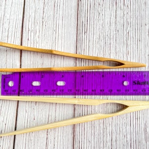 May include: Two pairs of natural bamboo tongs, one pair is 6 inches long and the other is 7 inches long, both are lying on a white wooden surface next to a purple ruler.