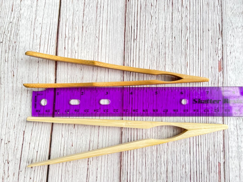 May include: Two pairs of natural bamboo tongs, one pair is 6 inches long and the other is 7 inches long, both are lying on a white wooden surface next to a purple ruler.