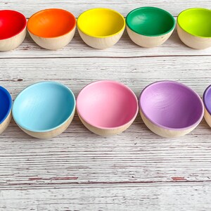 May include: Ten wooden bowls in a rainbow of colors, arranged in a row on a white wooden surface. The bowls are all the same size and shape, and they are painted in bright, vibrant colors.