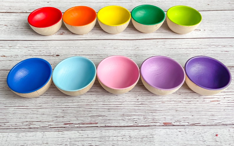 May include: Ten wooden bowls in a rainbow of colors, arranged in a row on a white wooden surface. The bowls are all the same size and shape, and they are painted in bright, vibrant colors.
