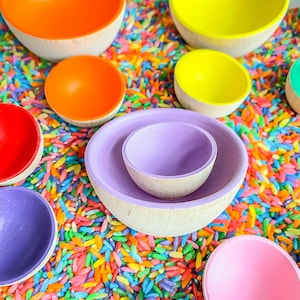 May include: A set of colorful wooden nesting bowls in rainbow colors, including red, orange, yellow, green, blue, purple, and pink. The bowls are arranged in a circle on a bed of colorful rice.