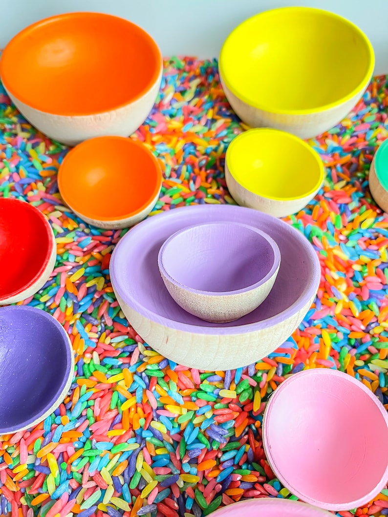 May include: A set of colorful wooden nesting bowls in rainbow colors, including red, orange, yellow, green, blue, purple, and pink. The bowls are arranged in a circle on a bed of colorful rice.