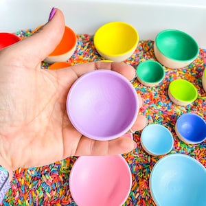 May include: A set of colorful wooden bowls in various sizes, nested inside each other. The bowls are painted in bright colors, including purple, pink, blue, green, yellow, and orange. The bowls are sitting in a tray filled with colorful sprinkles.