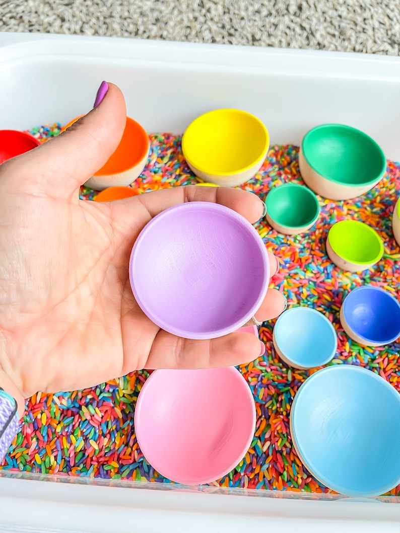May include: A set of colorful wooden bowls in various sizes, nested inside each other. The bowls are painted in bright colors, including purple, pink, blue, green, yellow, and orange. The bowls are sitting in a tray filled with colorful sprinkles.