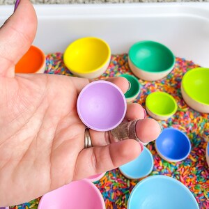 May include: A set of colorful wooden bowls in various sizes, nested inside each other. The bowls are painted in shades of purple, pink, blue, green, yellow, and orange. The bowls are sitting on a bed of rainbow-colored rice.