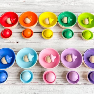 May include: A rainbow stacking toy set with wooden acorn-shaped tops and colorful bowls. The bowls are arranged in a rainbow pattern from red to purple. Each bowl has a smaller matching ball.