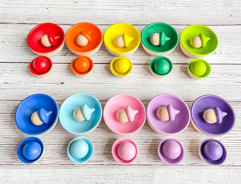 May include: A rainbow stacking toy set with wooden acorn-shaped tops and colorful bowls. The bowls are arranged in a rainbow pattern from red to purple. Each bowl has a smaller matching ball.