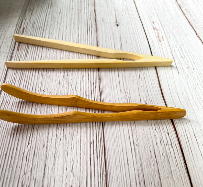 May include: Two pairs of wooden kitchen tongs on a white wooden surface. The tongs are made of natural bamboo and are ideal for serving food.