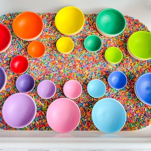 May include: A rainbow of colorful wooden bowls are nestled in a tray of rainbow sprinkles. The bowls are arranged in a circle, with the largest bowl in the center and the smallest bowls on the outside.