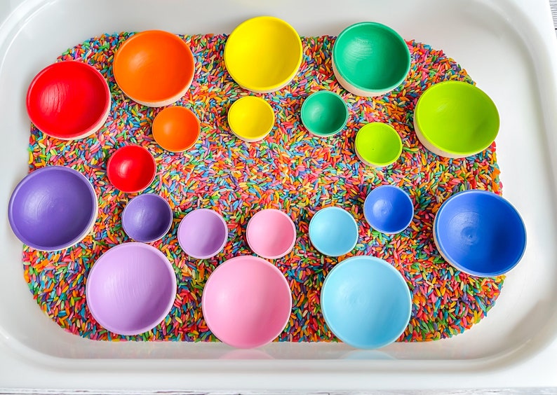 May include: A rainbow of colorful wooden bowls are nestled in a tray of rainbow sprinkles. The bowls are arranged in a circle, with the largest bowl in the center and the smallest bowls on the outside.