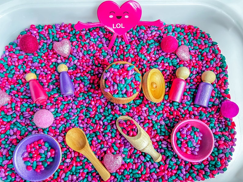 May include: A sensory bin filled with pink and green beans. There are wooden figurines, small bowls, a wooden spoon, and a pink heart with the word 'LOL' on it.