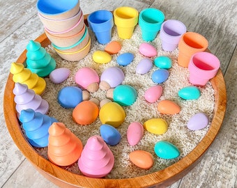 May include: A wooden bowl filled with white rice and colorful wooden eggs and cups. The bowl is surrounded by a variety of pastel-colored wooden cups and small trees.