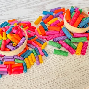 Op de afbeelding: Een verzameling kleurrijke pastabuizen in verschillende tinten rood, oranje, geel, groen, blauw en paars. De pasta is gerangschikt in drie stapels, met twee stapels in houten kommen en een stapel verspreid over een wit houten oppervlak.
