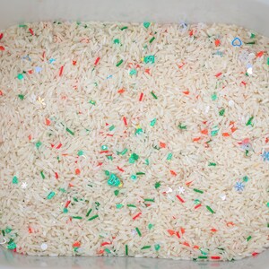 Puede incluir: Un recipiente de plástico blanco lleno de arroz blanco, decorado con chispas rojas, verdes y plateadas y pequeños confeti con forma de copo de nieve, corazón y estrella. Una actividad sensorial para niños.