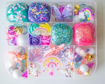 Op de afbeelding: Een doorzichtige plastic organizer gevuld met kleurrijke knutselspullen. De vakken bevatten glinsterende slijm, decoratieve bloemen, vlinders, regenbogen, eenhoorns en andere versieringen in roze, paars, blauw en geel.