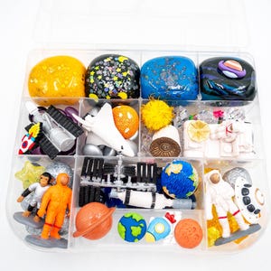 May include: A clear plastic storage container filled with space-themed sensory items, including four blocks of colored slime, miniature plastic astronauts, a space shuttle, a rocket, a globe, planets, and various small space-related figurines.