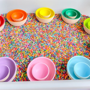 May include: A rainbow of wooden nesting bowls are arranged in a circle on a bed of colorful rice. The bowls are all different sizes and are made of natural wood.