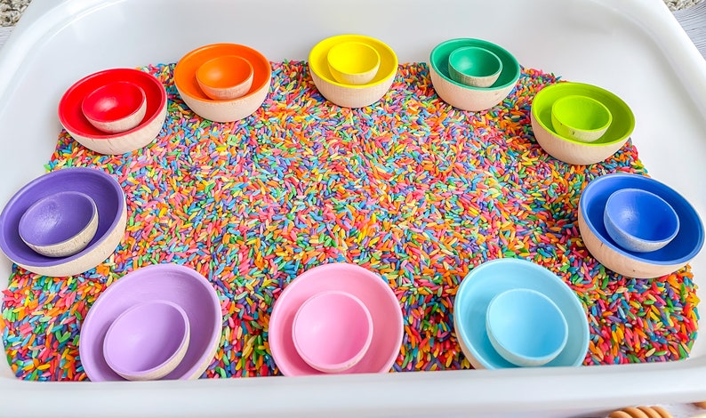 May include: A rainbow of wooden nesting bowls are arranged in a circle on a bed of colorful rice. The bowls are all different sizes and are made of natural wood.