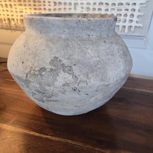 May include: A weathered, gray ceramic pot with a rounded body and a wide, slightly flared rim. The surface shows signs of age and texture, with darker patches adding visual interest. The pot sits on a dark wooden surface.