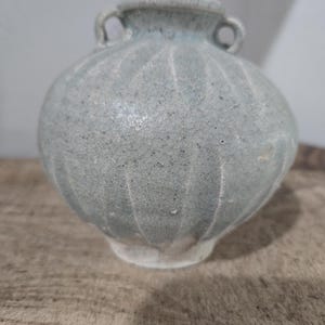 May include: A small, round, light blue ceramic vase with a textured surface and two small handles. The vase has vertical ridges and a slightly flared rim. The base is a lighter color, and the vase sits on a wooden surface.