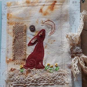 May include: Handmade fabric art piece featuring a red rabbit silhouette gazing at a crescent moon. The artwork includes a button, embroidered flowers, and a burlap accent. The piece is on a textured, stained fabric with a lace trim.