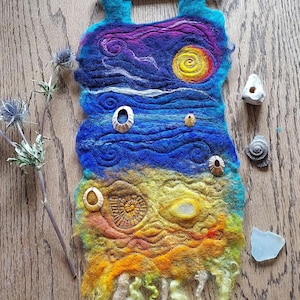 Puede incluir: Una pared colgante de fieltro colorida que representa una puesta de sol sobre el océano. La pared colgante está compuesta por cuatro capas de fieltro, cada una con un color y una textura diferentes. La capa superior es un fieltro azul oscuro con una puesta de sol púrpura y blanca. La segunda capa es un fieltro azul claro con olas blancas. La tercera capa es un fieltro amarillo con una playa de arena marrón. La capa inferior es un fieltro amarillo con una franja. La pared colgante está decorada con pequeñas conchas.