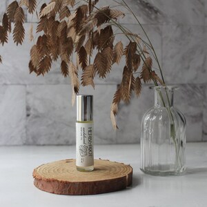 May include: A roll-on perfume oil bottle with a silver cap and a label that reads "HERB & ROOT". The bottle sits on a wooden slice, with dried foliage and a clear glass vase in the background. The perfume oil is labeled "sandalwood".