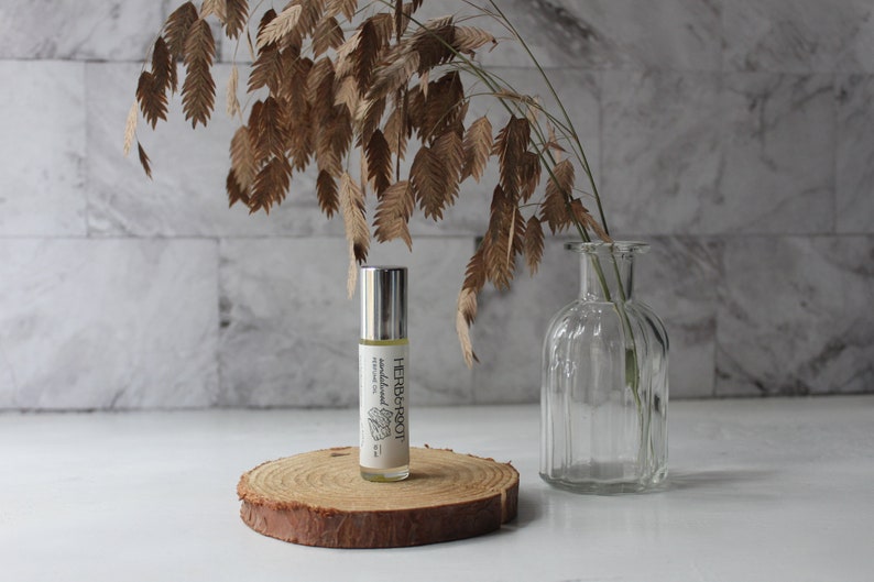 May include: A roll-on perfume oil bottle with a silver cap and a label that reads "HERB & ROOT". The bottle sits on a wooden slice, with dried foliage and a clear glass vase in the background. The perfume oil is labeled "sandalwood".