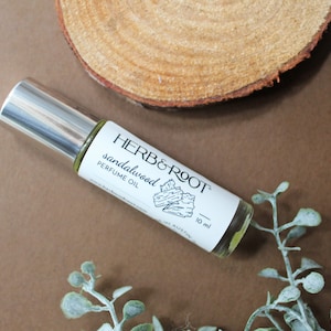 May include: A clear glass roll-on bottle of sandalwood perfume oil with a silver cap, labeled "HERB & ROOT." The bottle is 10 ml. The background includes a wooden slice and greenery on a brown surface.