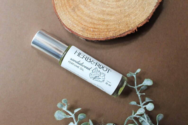 May include: A clear glass roll-on bottle of sandalwood perfume oil with a silver cap, labeled "HERB & ROOT." The bottle is 10 ml. The background includes a wooden slice and greenery on a brown surface.