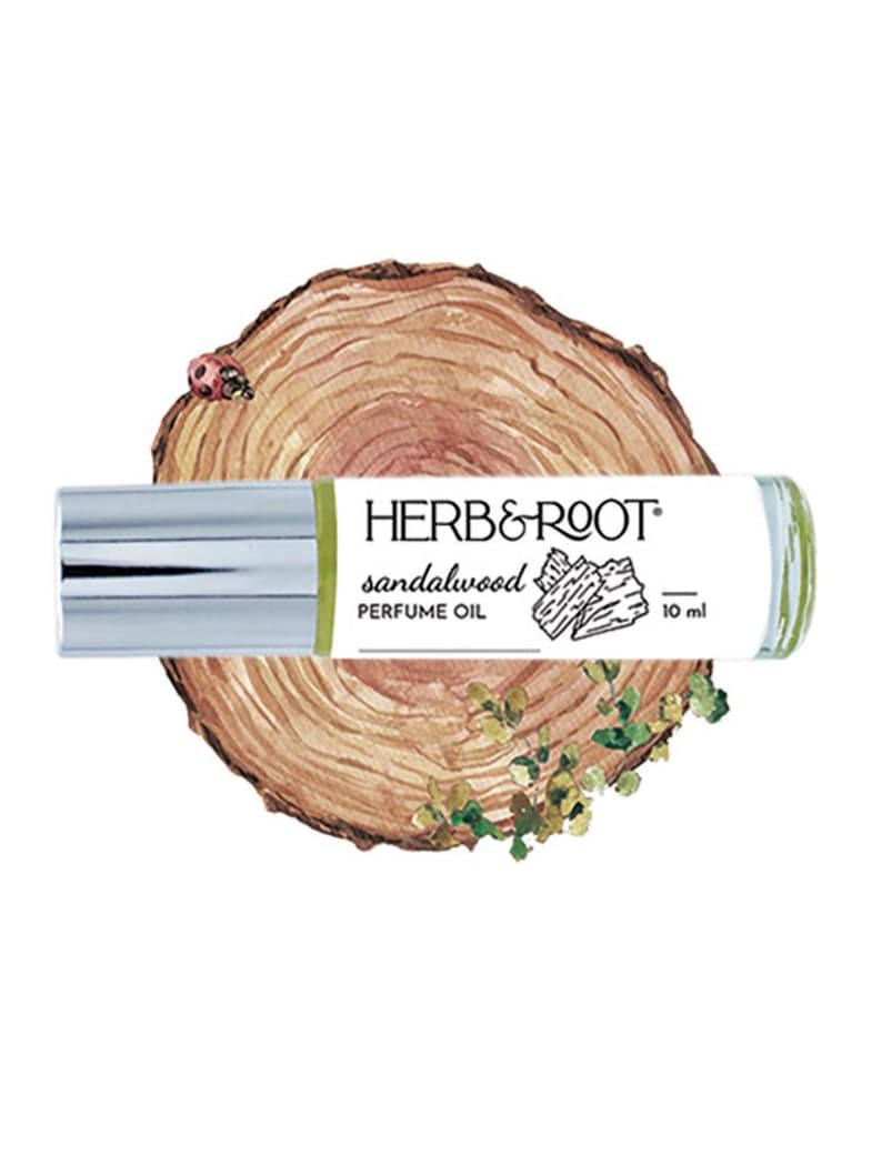 May include: A sandalwood perfume oil roll-on bottle with a silver cap, labeled "HERB & ROOT." The bottle is set against a backdrop of a wood slice with a ladybug and greenery. The bottle contains 10 ml of perfume.