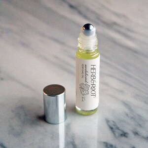 May include: A small, clear glass roll-on bottle of sandalwood perfume oil with a silver cap. The label reads "HERB & ROOT" and "sandalwood perfume oil". The bottle is 10 ml. The cap is next to the bottle.
