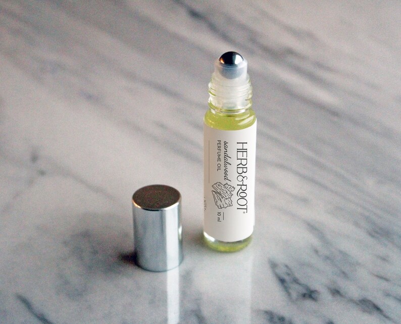 May include: A small, clear glass roll-on bottle of sandalwood perfume oil with a silver cap. The label reads "HERB & ROOT" and "sandalwood perfume oil". The bottle is 10 ml. The cap is next to the bottle.
