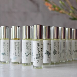 May include: A collection of roll-on perfume bottles with silver caps and white labels. The labels feature the brand name "HERB & ROOT" and various botanical illustrations. The bottles are arranged in a semi-circle, with a blurred background of a vase and flowers.