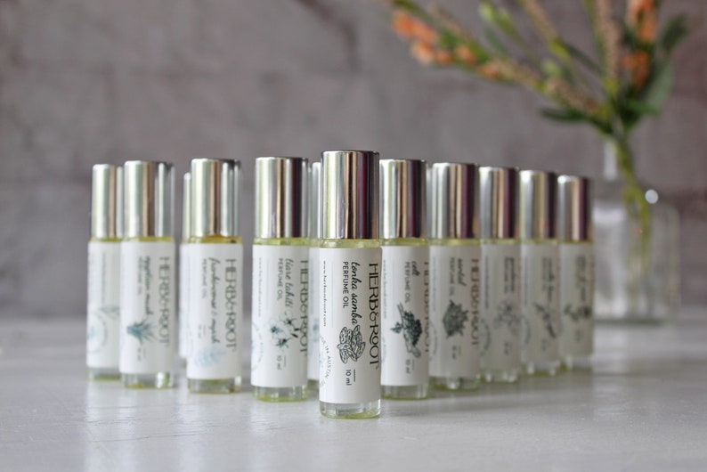 May include: A collection of roll-on perfume bottles with silver caps and white labels. The labels feature the brand name "HERB & ROOT" and various botanical illustrations. The bottles are arranged in a semi-circle, with a blurred background of a vase and flowers.