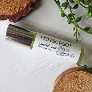 May include: A roll-on perfume oil bottle with a silver cap and a white label that reads "HERB & ROOT sandalwood PERFUME OIL." The bottle is 10 ml. The background includes wood slices and green foliage.