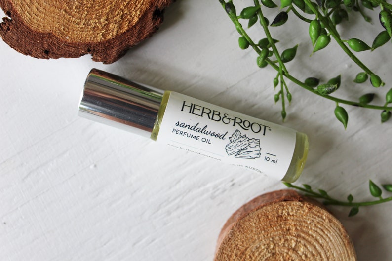 May include: A roll-on perfume oil bottle with a silver cap and a white label that reads "HERB & ROOT sandalwood PERFUME OIL." The bottle is 10 ml. The background includes wood slices and green foliage.