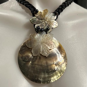 May include: A black braided cord necklace with a large, round, iridescent shell pendant. The pendant has a smaller, carved shell flower on top.