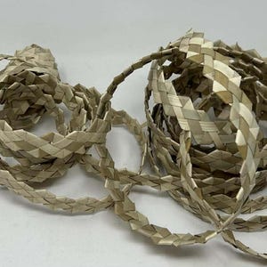 Puede incluir: Un rollo enrollado de cinta tejida de color beige claro. La cinta está hecha de finas tiras planas de material, intrincadamente tejidas para crear una apariencia trenzada texturizada. La cinta es de color neutro.