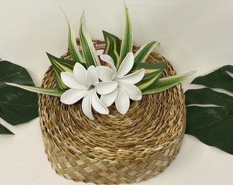 May include: A woven straw plate with white plumeria flowers and green and white variegated leaves. The plate is round and has a natural, rustic look.