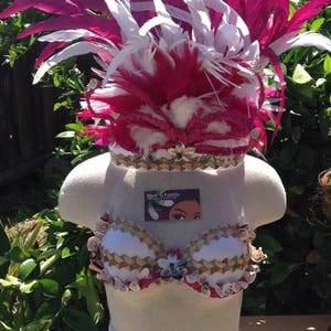 May include: A white mannequin torso wearing a white and brown bra with pink and white feathers on top of its head. The bra is decorated with seashells and the feathers are arranged in a mohawk style. The mannequin is standing on a wooden surface. The text "Bare O' Pasifika" is visible on a business card.