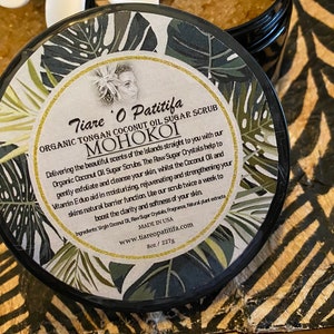 K&ouml;nnte beinhalten: Ein schwarzer Topf mit goldenem Deckel und einem wei&szlig;en Etikett mit gr&uuml;nen tropischen Bl&auml;ttern. Das Etikett lautet "Tiare O Patitifa Organic Tongan Coconut Oil Sugar Scrub MOHOKOI". Der Text beschreibt die Vorteile und Inhaltsstoffe des Produkts.