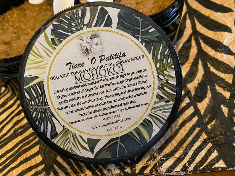 K&ouml;nnte beinhalten: Ein schwarzer Topf mit goldenem Deckel und einem wei&szlig;en Etikett mit gr&uuml;nen tropischen Bl&auml;ttern. Das Etikett lautet "Tiare O Patitifa Organic Tongan Coconut Oil Sugar Scrub MOHOKOI". Der Text beschreibt die Vorteile und Inhaltsstoffe des Produkts.