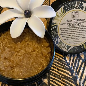 K&ouml;nnte beinhalten: Ein schwarzer Topf gef&uuml;llt mit einem braunen Zuckerschrub. Der Topf hat eine wei&szlig;e Blume oben und ein Etikett mit der Aufschrift "Tiare O Patitifa Organic Tongan Coconut Oil Sugar Scrub MOHOKOI".